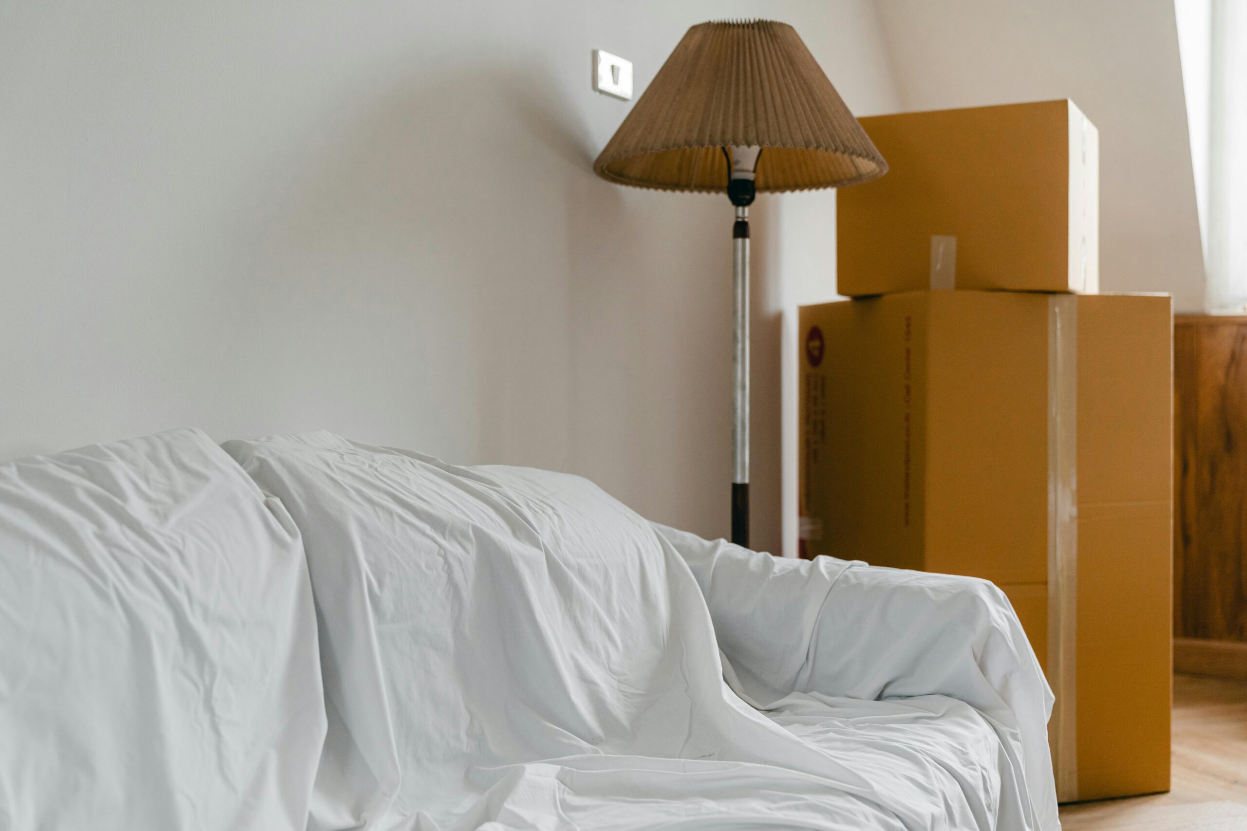 Wohnungsauflösung - Ablauf und wichtige Tipps Covered with sheet sofa and floor lamp and packed cardboard boxes in light spacious room of new contemporary house