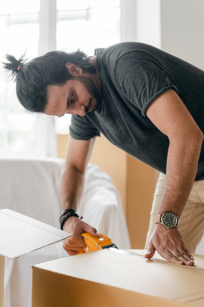 A man carefully sealing moving boxes with a tape dispenser, preparing for relocation.