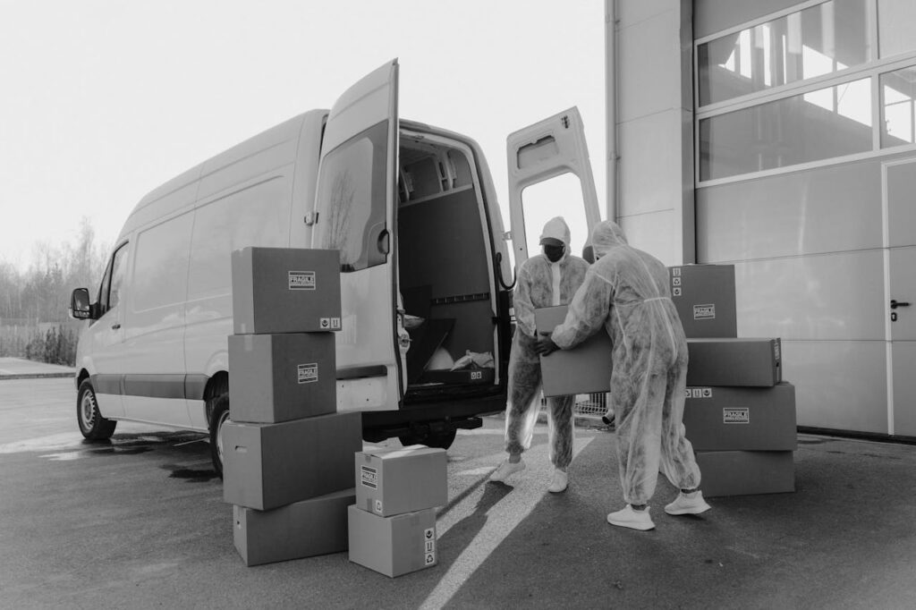 Two workers in PPE handling packages from a delivery van in a monochrome setting.