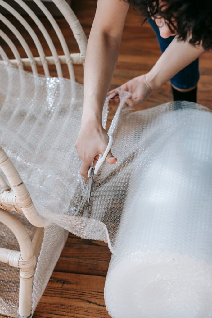 A woman using scissors to cut bubble wrap for packing a wicker chair indoors, preparing to move.